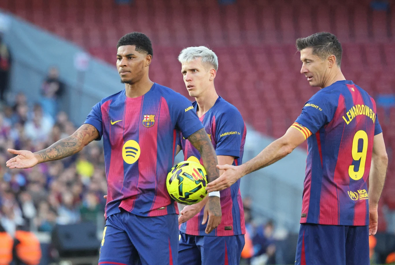 Trzech piłkarzy w strojach FC Barcelony na boisku, jeden z nich trzyma piłkę, a zawodnik z numerem 9 i napisem 'Lewandowski' na plecach wskazuje ręką kierunek gry; w tle widoczna trybuna stadionu.