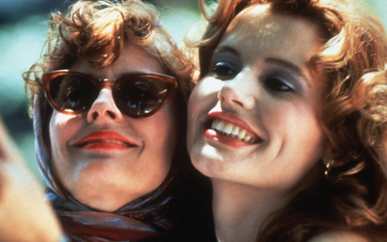 Dwie kobiety uśmiechnięte i blisko siebie, jedna z nich nosi okulary przeciwsłoneczne i apaszkę na głowie, druga ma rozpuszczone włosy i mocno podkreślone oczy makijażem, obie wyglądają na zadowolone i szczęśliwe.