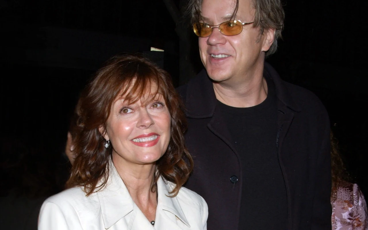 kobieta w białej marynarce uśmiecha się, stojąc obok mężczyzny w ciemnych okularach i czarnej kurtce; oboje spoglądają w różnych kierunkach, a tło jest zacienione