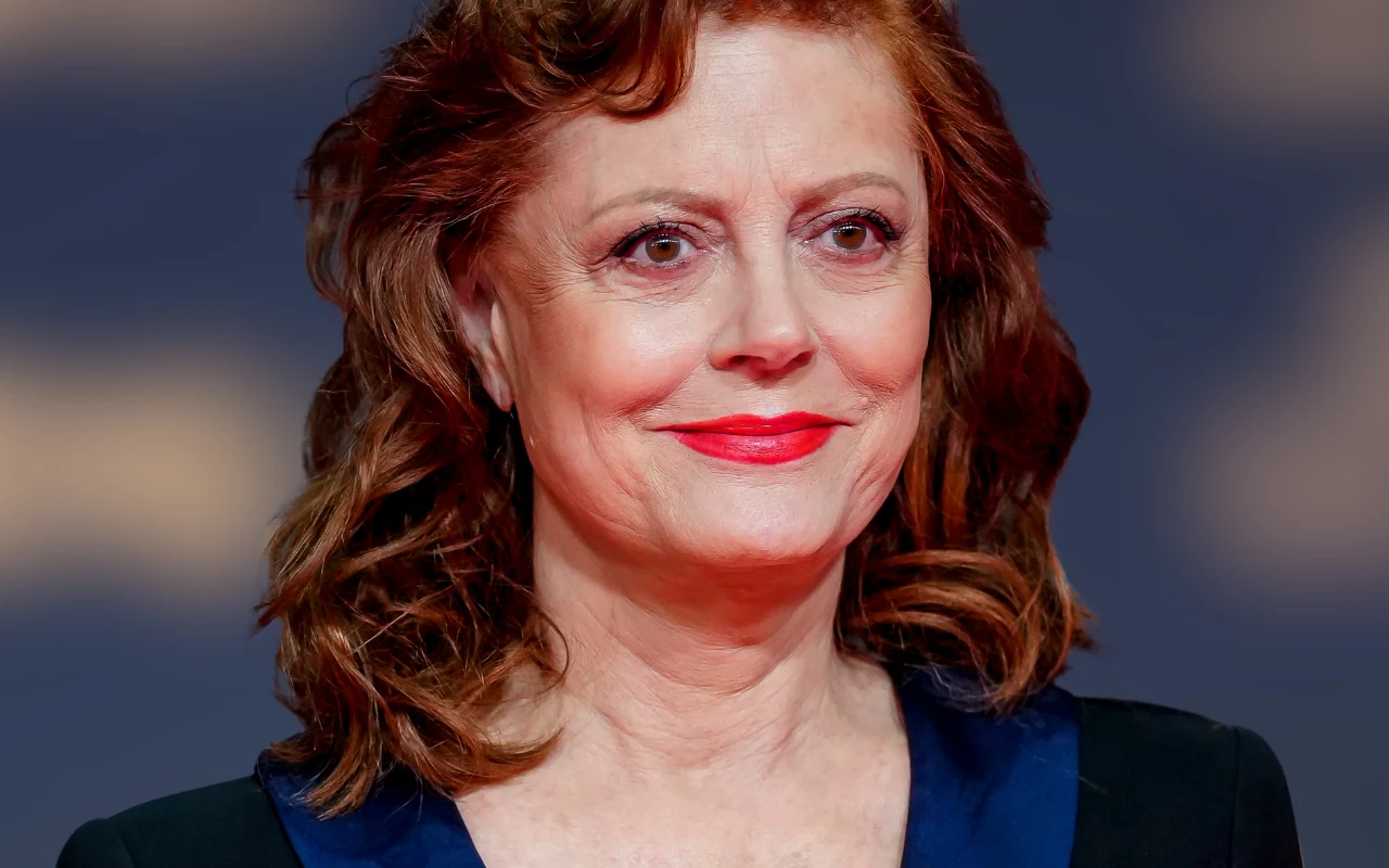 Kobieta w średnim wieku z falowanymi, rudobrązowymi włosami i wyrazistym makijażem, uśmiechnięta, w eleganckiej czarnej marynarce z niebieskimi klapami na tle rozmytych, jasnych plam.