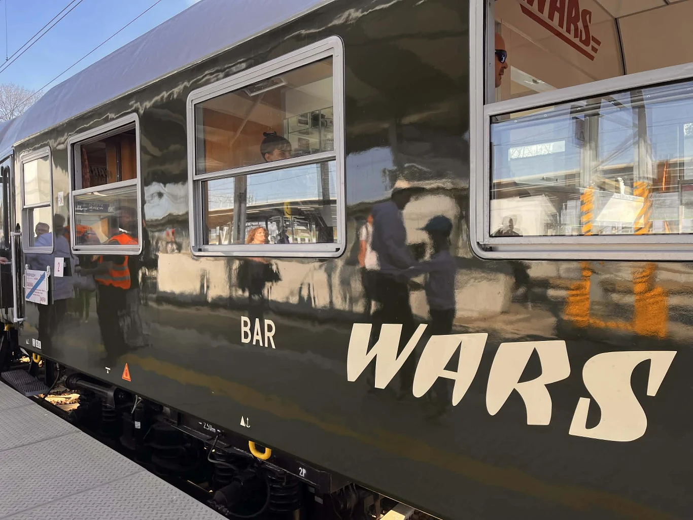 Wagony z zewnątrz prezentują się elegancko wagon restauracyjny pociągu z dużym białym napisem 'BAR WARS' na ciemnozielonej burcie, przez okna widoczni są pasażerowie i obsługa, w odbiciu na lakierze wyraźnie rysują się sylwetki ludzi stojących na peronie