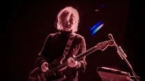 Muzyk z długimi jasnymi włosami grający na gitarze elektrycznej, oświetlony scenicznym światłem, skupiony na grze na tle ciemnej sceny z widocznym statywem mikrofonowym i pulpitem z nutami obok.