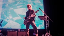 Muzyk grający na gitarze elektrycznej stoi na scenie oświetlonej światłem fioletowo-niebieskim, w tle widoczna jest duża projekcja przedstawiająca z lotu ptaka krajobraz pól, obok muzyka stoją wzmacniacze, mikrofon oraz płynna butelka na podłodze, a wo...