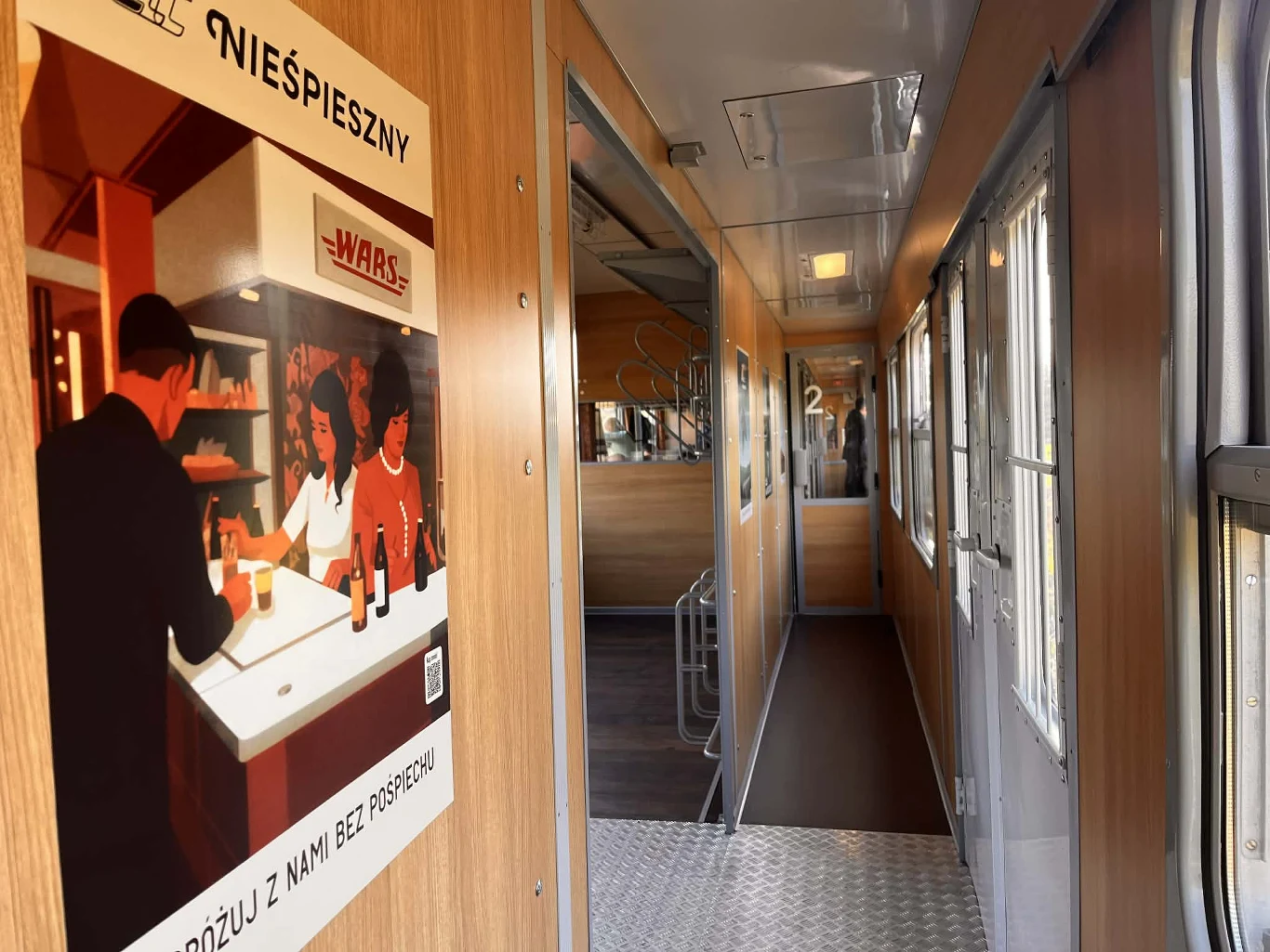 Nieśpiesznym można objechać całą Polskę wnętrze wagonu pociągu z jasnymi panelami ściennymi, metalową podłogą w korytarzu oraz plakatem promującym bar WARS, na którym widać rysunkowe postaci przy ladzie