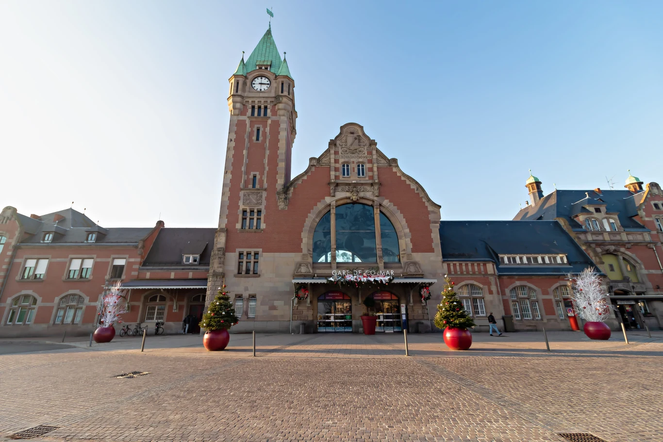 Dworzec w Colmar wygląda znajomo, prawda? zabytkowy budynek dworca kolejowego z wysoką wieżą zegarową, duże ozdobne okno nad wejściem głównym, przed wejściem ustawiono donice z choinkami ozdobionymi świątecznymi dekoracjami