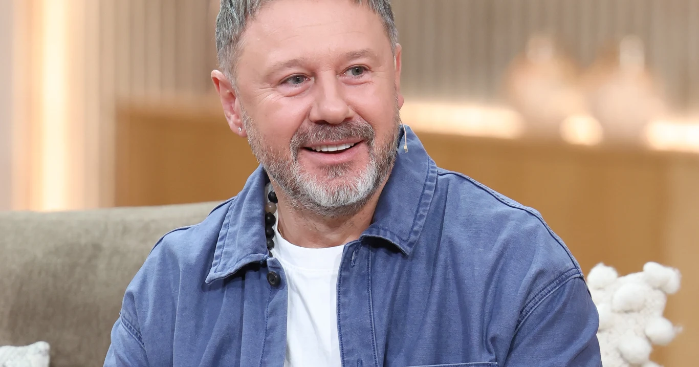 Andrzej Piaseczny Andrzej Piaseczny w jasnej koszuli i białym t-shircie siedzi na beżowej sofie w przytulnym wnętrzu.
