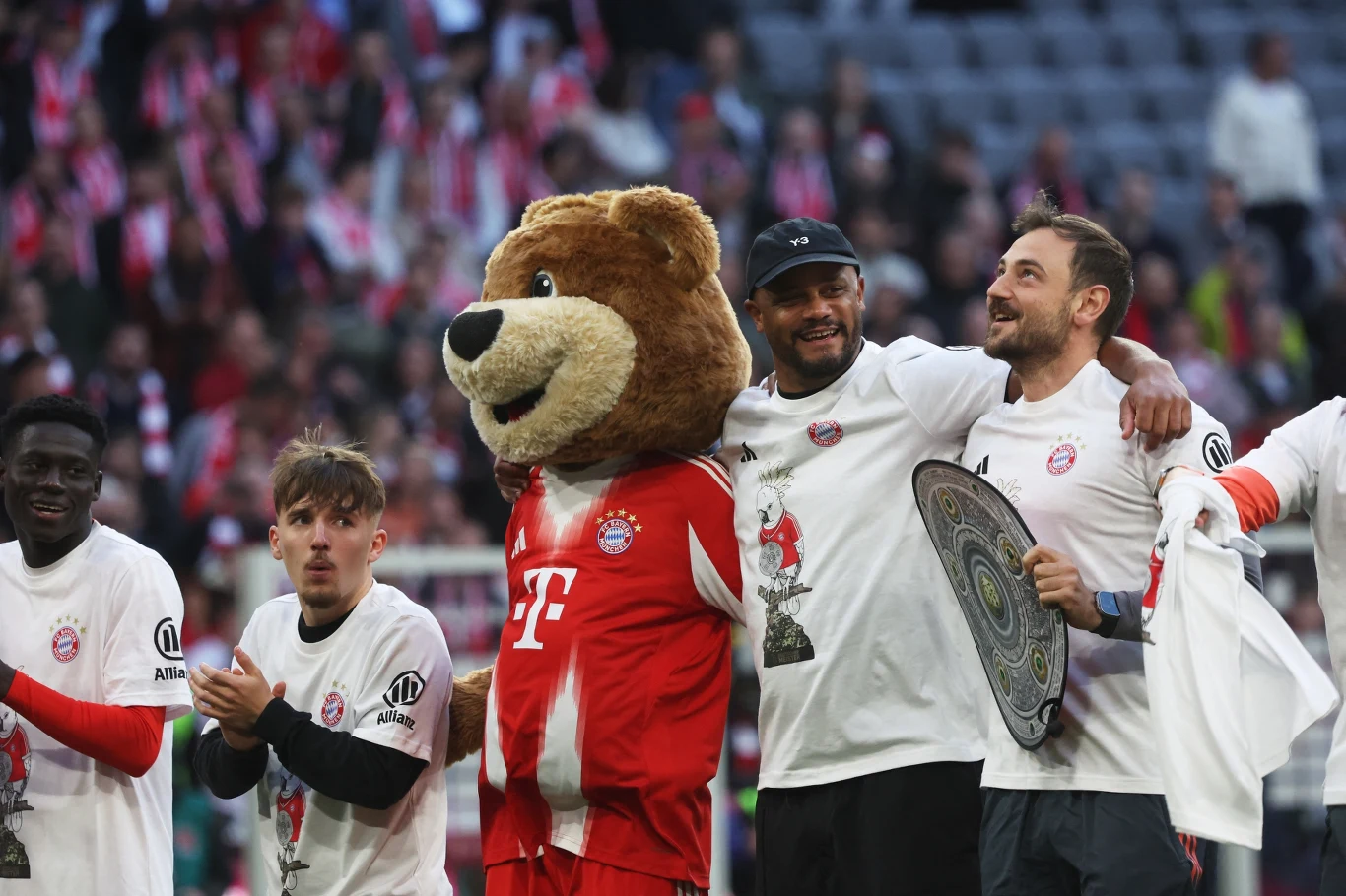 piłkarze w białych koszulkach świętują na stadionie, maskotka drużyny w stroju Bayernu Monachium stoi pośród nich, jeden z zawodników trzyma trofeum, wszyscy uśmiechnięci w otoczeniu kibiców