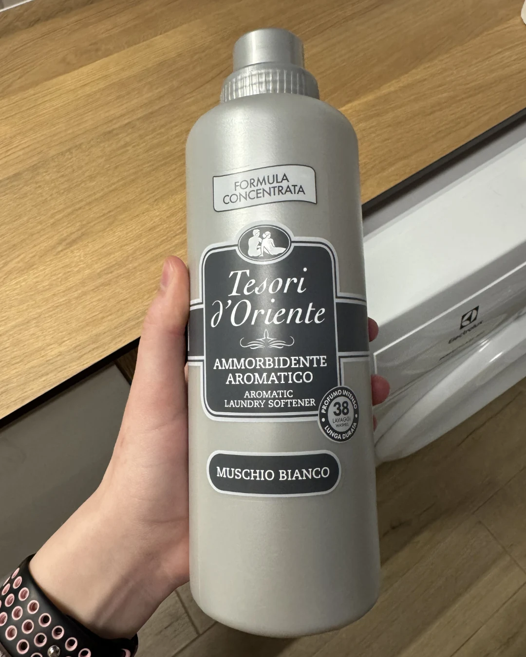 Ten jeden zapach wygrał u mnie wszystko Butelka szarego płynu do zmiękczania tkanin Tesori d'Oriente o zapachu białego piżma trzymana w lewej ręce na tle drewnianego blatu i białej pralki.