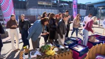 Grupa osób ogląda stoiska z żywnością oraz różnymi produktami wystawionymi w pojemnikach na świeżym powietrzu, pośród których znajdują się m.in. banany, kubki, warzywa oraz książki; uczestnicy zainteresowani są ofertą, niektórzy robią zdjęcia telefonami.