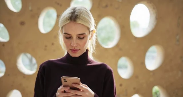 Kobieta z blond włosami ubrana w ciemny golf skupia się na smartfonie, stojąc przed jasną ścianą z dużymi, nieregularnymi otworami przepuszczającymi światło.