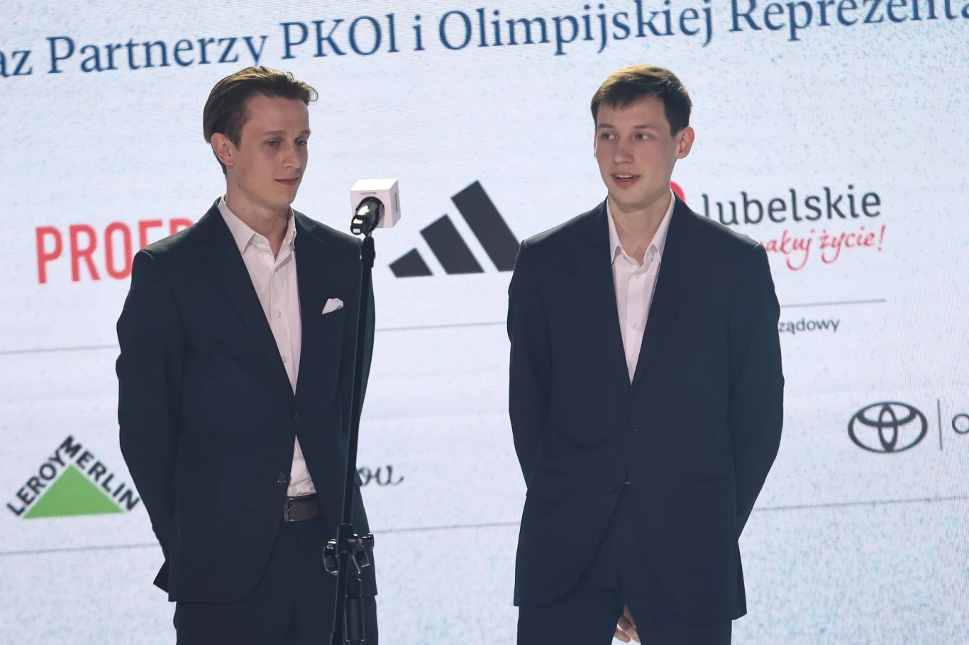 Paweł Wąsek i Kacper Tomasiak na gali PKOl Dwóch młodych mężczyzn w garniturach stoi obok siebie przed mikrofonem na tle ekranu z logotypami znanych marek, w tym Adidas i Toyota, w formalnym otoczeniu eventowym.