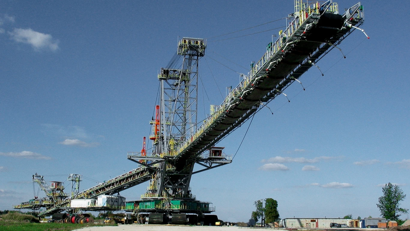 Zwałowarka ZGOT 15400 ma ogromne rozmiary. Ogromna koparka wielonaczyniowa z długim wysięgnikiem i licznymi elementami metalowymi, osadzona na gąsienicach, dominuje w krajobrazie przemysłowym; w tle widoczne są niewielkie zabudowania oraz drzewa pod jasnym niebem.
