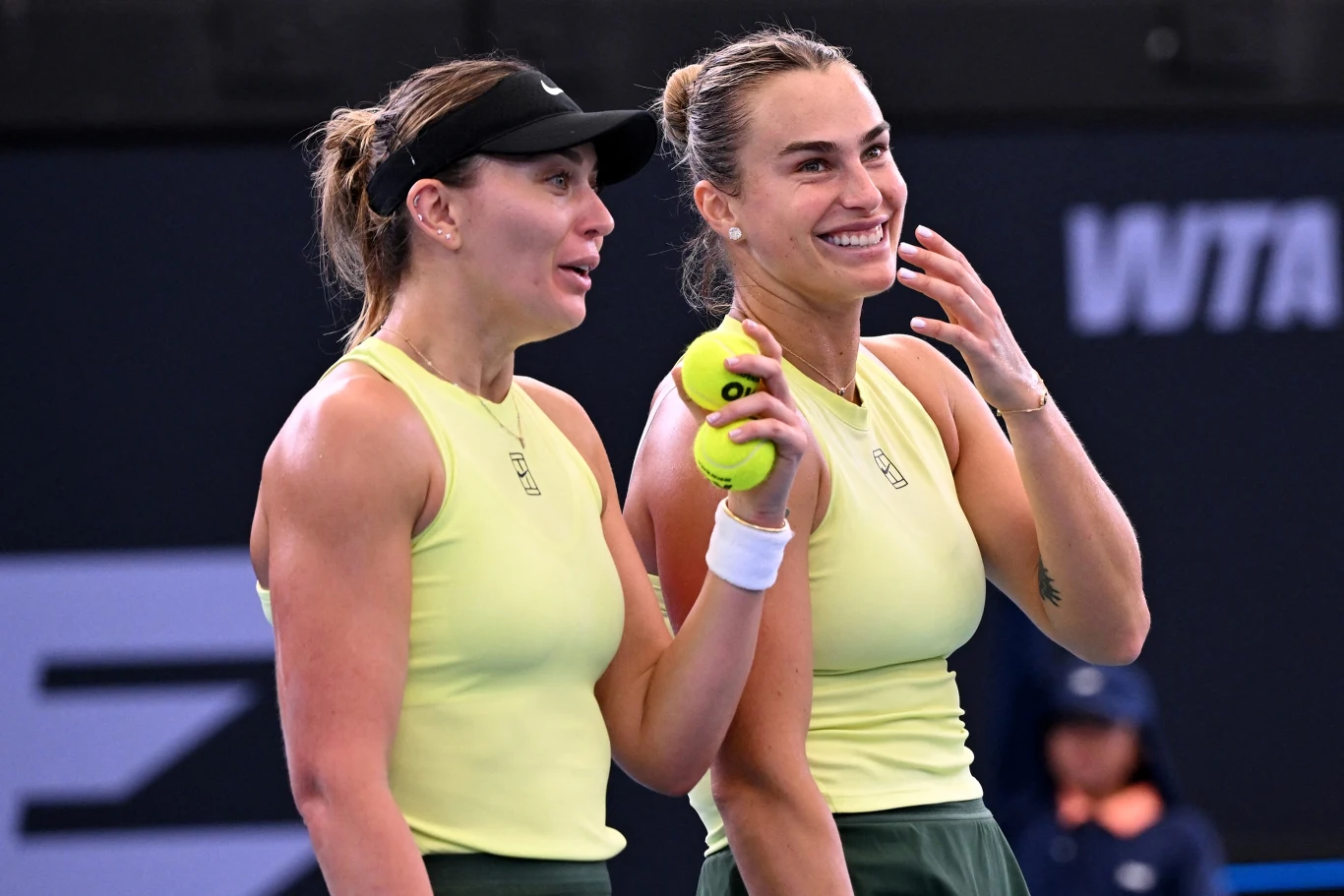 Paula Badosa i Aryna Sabalenka Dwie tenisistki w żółtych strojach rozmawiają i uśmiechają się na korcie, jedna trzyma piłki tenisowe w dłoni, w tle rozmyta postać sędzi oraz elementy boiska.