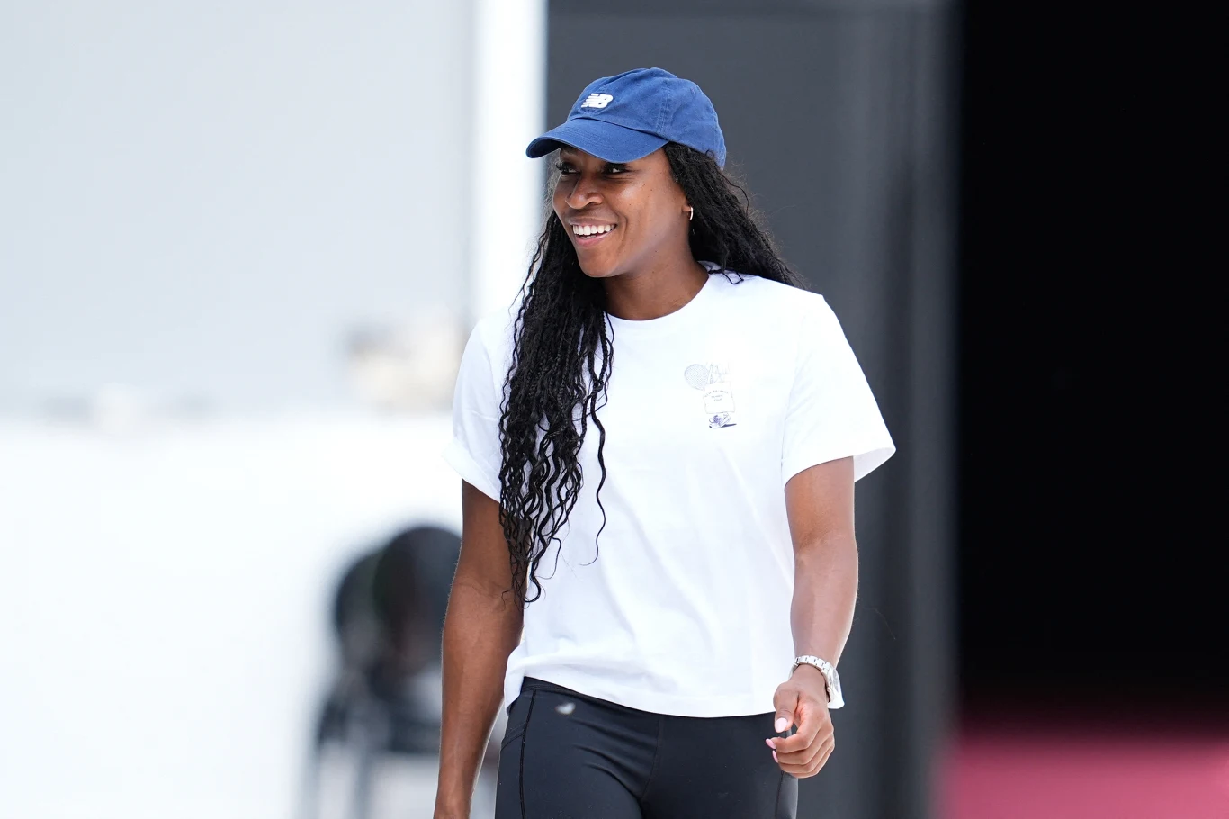 Coco Gauff Kobieta o długich, kręconych włosach ubrana w białą koszulkę, czapkę z daszkiem i czarne legginsy uśmiecha się, spacerując po jasnym tle z niewyraźnymi ciemnymi elementami w tle.