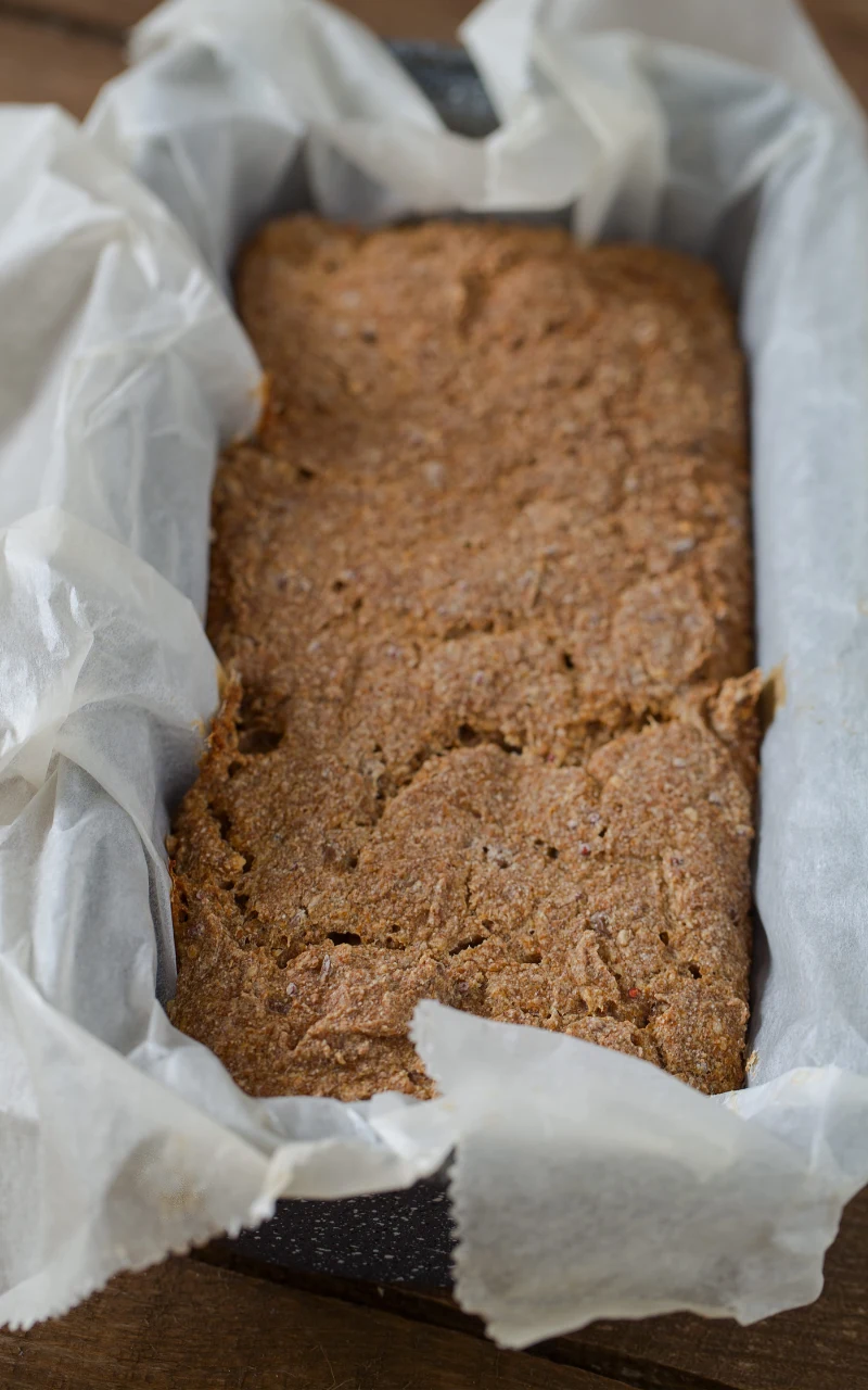 Bochenek domowego chleba razowego na zakwasie leży w foremce wyłożonej papierem do pieczenia, powierzchnia wypieku jest nierówna i lekko popękana.