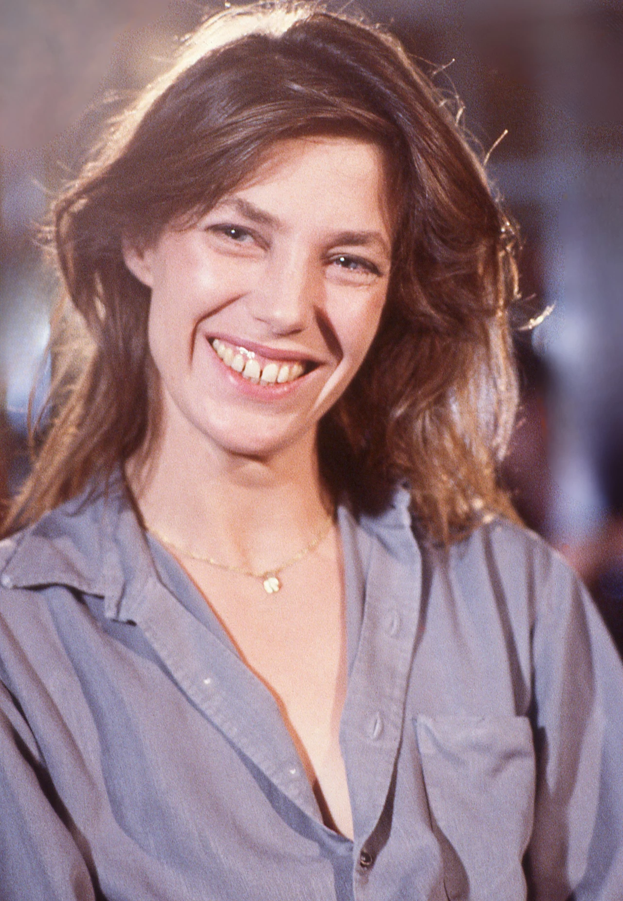 Uśmiechnięta kobieta o długich, rozpuszczonych włosach, ubrana w luźną, jasnoszarą koszulę oraz delikatny złoty naszyjnik z zawieszką, patrzy prosto w obiektyw na rozświetlonym tle.