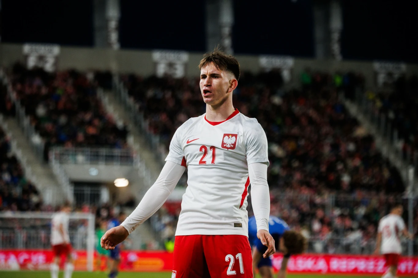 Marcel Reguła piłkarz w biało-czerwonym stroju reprezentacji Polski z numerem 21 stoi na murawie stadionu podczas meczu, z widocznymi emocjami na twarzy, za nim rozmyte sylwetki innych zawodników oraz wypełnione trybuny