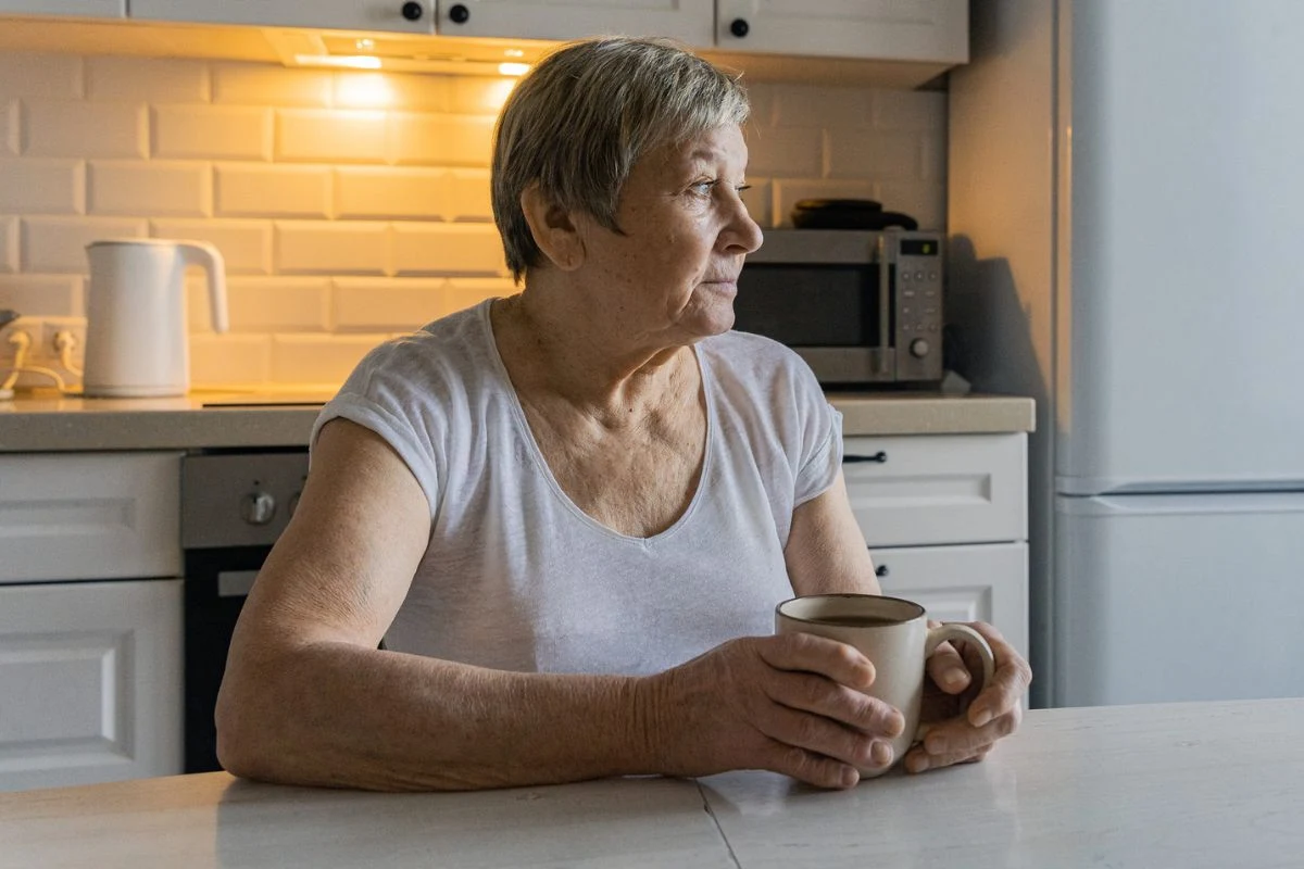Zawał mięśnia sercowego może inaczej objawiać się u kobiet. Często przypomina zatrucie pokarmowe, z towarzyszącymi nudnościami, wymiotami i bólami brzucha Starsza kobieta z siwymi włosami siedzi przy stole w kuchni, trzyma kubek; w tle czajnik i mikrofalówka.