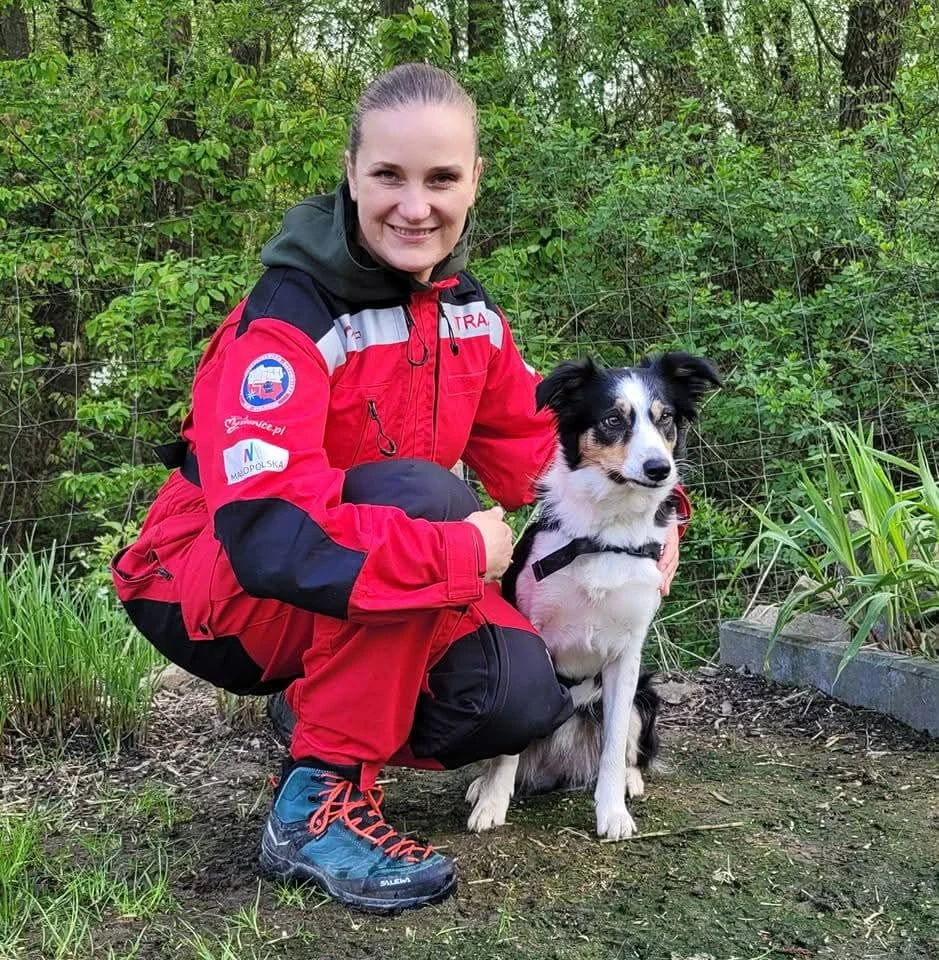 Katarzyna Harmata od wielu lat pracuje z psami w różnych obszarach życia społecznego kobieta w czerwonym kombinezonie ratowniczym kuca obok czarno-białego psa w uprzęży, oboje znajdują się w zielonym, leśnym otoczeniu, na twarzach widać spokój i gotowość do działania