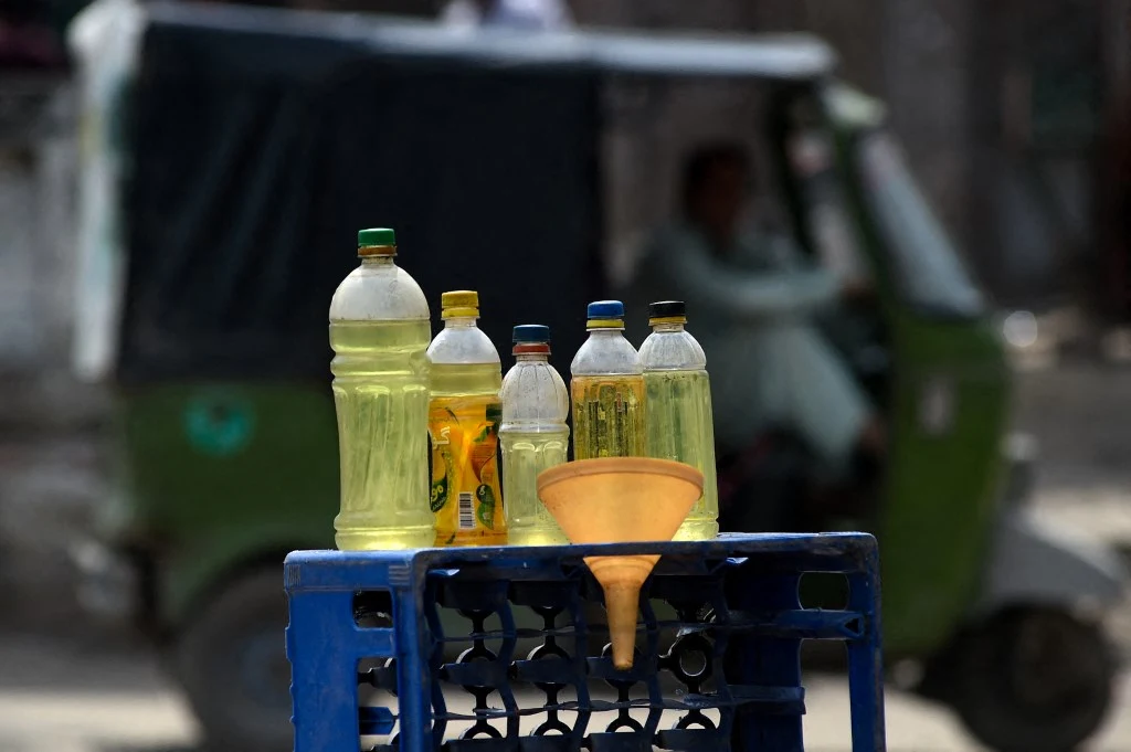 Butelki z benzyną przy drodze w Lahore w Pakistanie, 25 marca 2026 r. plastikowe butelki wypełnione żółtym płynem ustawione na niebieskiej skrzynce, obok żółty lejek; w tle rozmazany pojazd auto-riksza z kierowcą