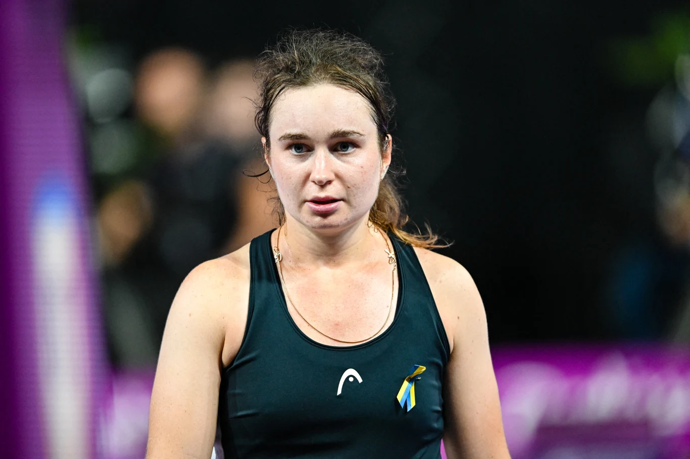 młoda kobieta w sportowym stroju stoi na korcie tenisowym, na jej stroju przypięta jest wstążka w kolorach niebieskim i żółtym, twarz napięta i skupiona, tło rozmazane i ciemne