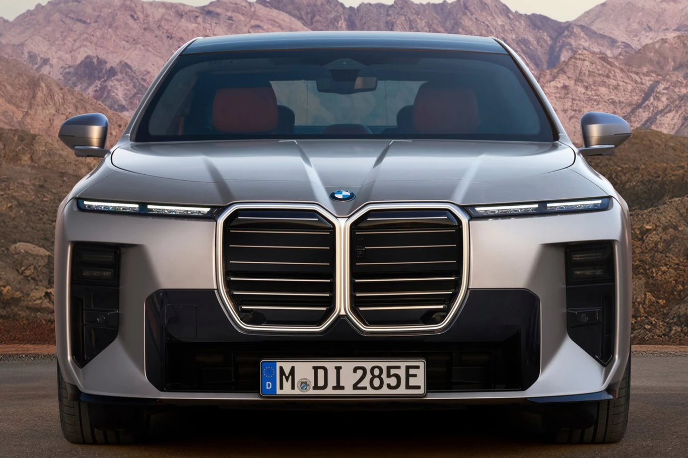 Zamiast podwójnych świateł głównych ukrytych pod prostokątnym kloszem, BMW serii 7 otrzymało kwadratowe reflektory optycznie zlewające się z czarnymi listwami Srebrny samochód BMW o muskularnej sylwetce z charakterystycznym, dużym grillem oraz wąskimi reflektorami LED na tle skalistego górskiego krajobrazu.