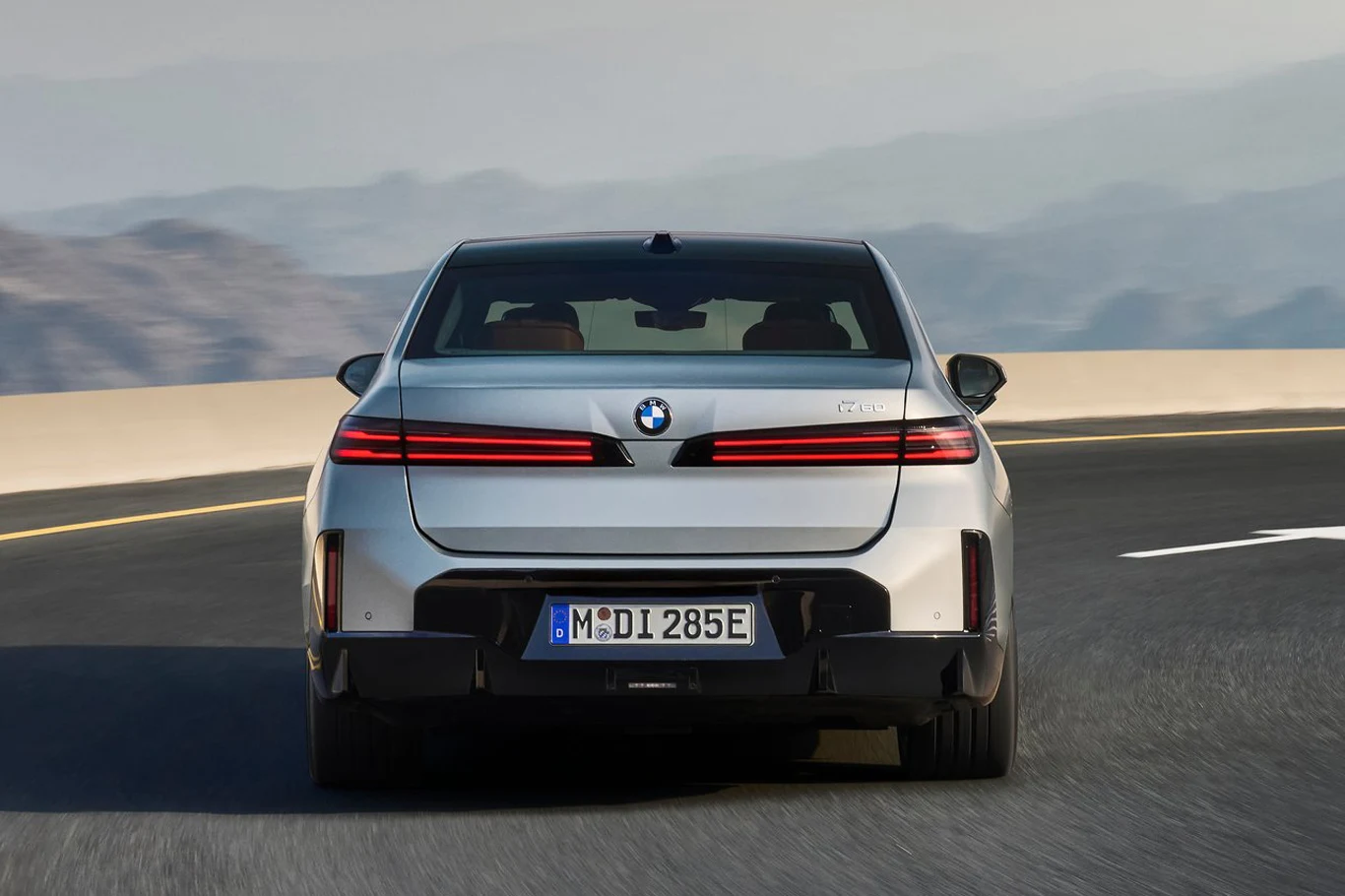 Tył nowego BMW serii 7 sprawia wrażenie, że mamy do czynienia z zupełnie nowym modelem Neue Klasse srebrne BMW z nowoczesnymi, wąskimi tylnymi światłami i niemiecką rejestracją jedzie autostradą na tle górskiego krajobrazu