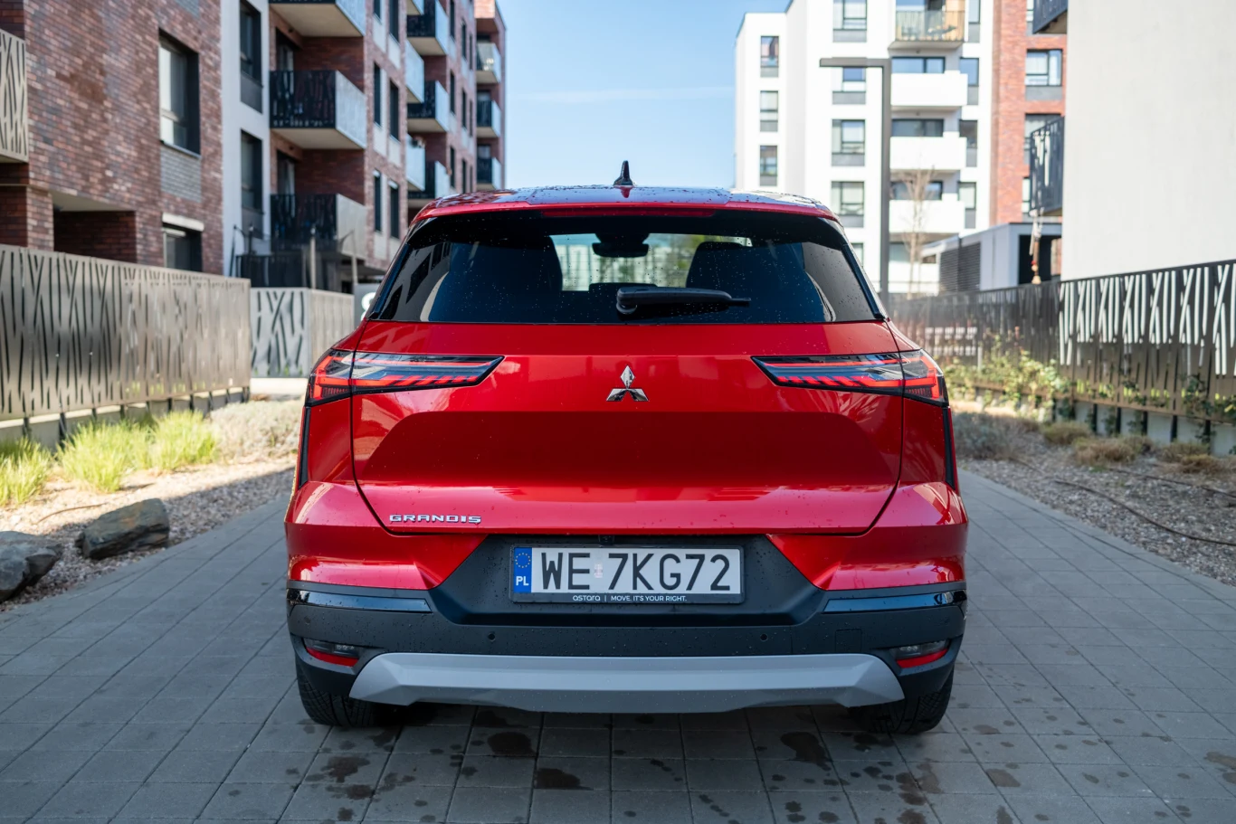 czerwony samochód Mitsubishi o nowoczesnym designie widoczny od tyłu, zaparkowany na tle miejskiej zabudowy z apartamentowcami po obu stronach wąskiej alejki