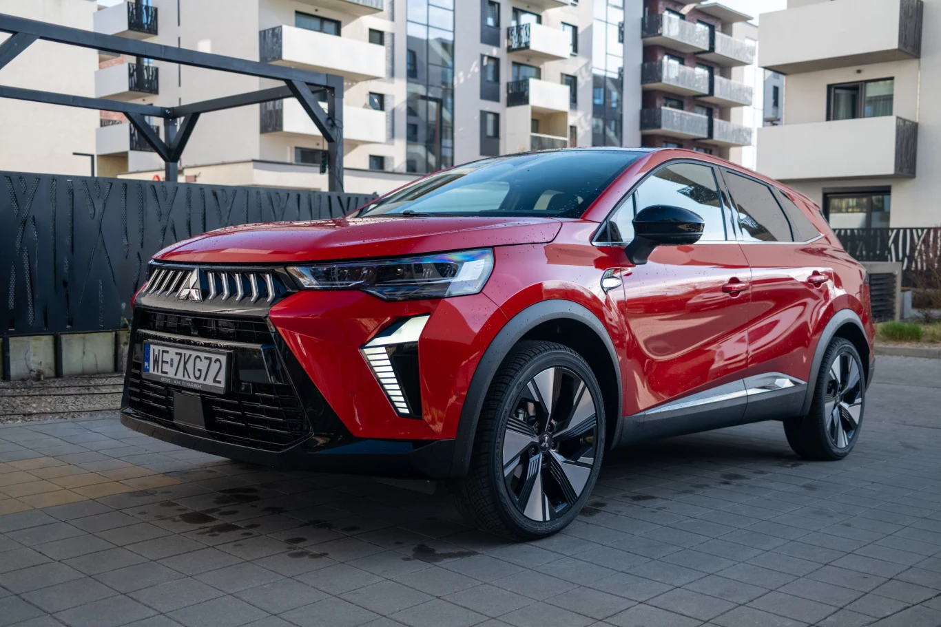 Czerwony nowoczesny SUV Mitsubishi z czarnymi elementami wykończenia stoi na parkingu przed blokami mieszkalnymi, odbijając w lakierze otoczenie i światło dzienne, wyróżniając się dużymi felgami oraz charakterystycznym grillem na froncie.