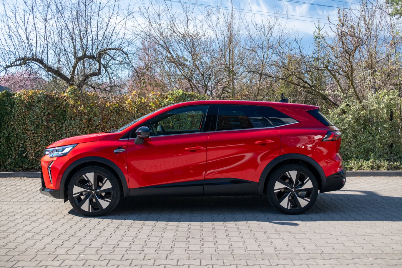 czerwony samochód elektryczny typu SUV zaparkowany na chodniku, nowoczesny design z dużymi felgami, czarne wykończenia wokół okien i dolnych partii nadwozia, w tle gęste krzewy i drzewa bez liści