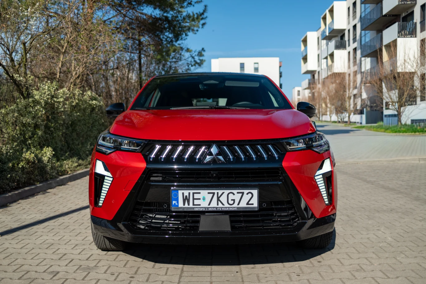 czerwony SUV Mitsubishi Eclipse Cross ustawiony na środku betonowej drogi, z wyraźnie widoczną tablicą rejestracyjną oraz nowoczesnym, agresywnym przednim pasem i charakterystycznymi światłami LED, w tle osiedlowe budynki mieszkalne i drzewa po lewej s...