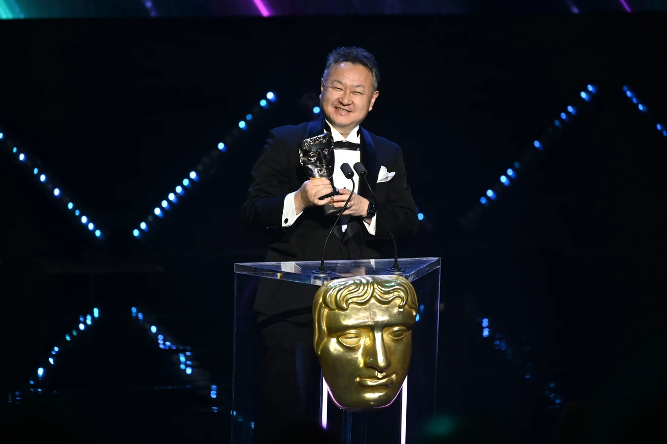 Shuhei Yoshida Shuhei Yoshida w smokingu ze statuetką przy mównicy z maską BAFTA, w tle kolorowe światła LED.