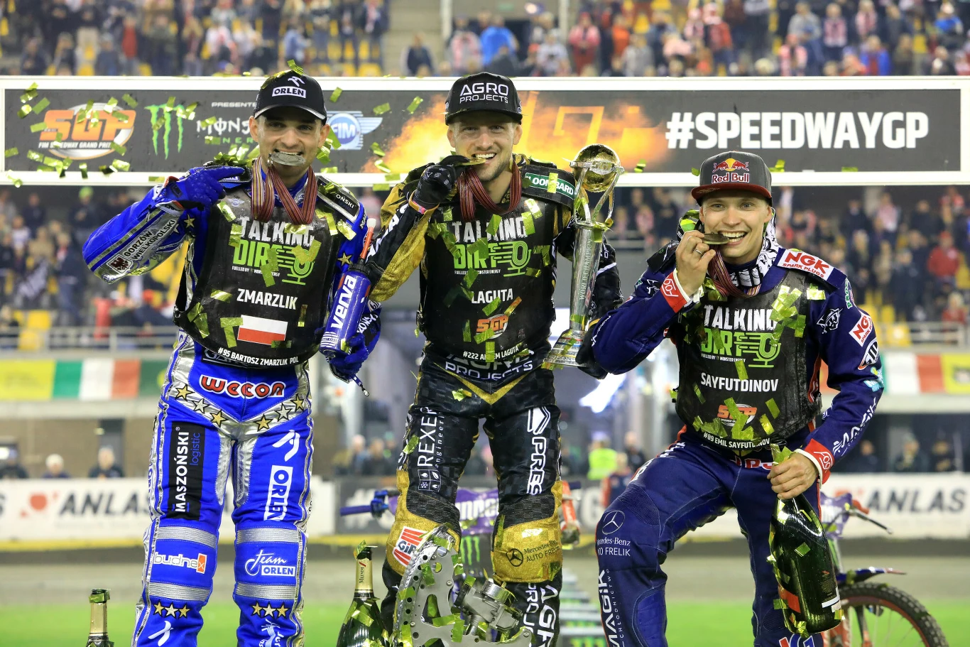 Trzech uśmiechniętych zawodników żużlowych w strojach sportowych stoi na podium, każdy z medalem na szyi, środkowy trzyma w ręce puchar, w tle baner z napisem #SPEEDWAYGP oraz podekscytowana publiczność w oświetlonym stadionie.