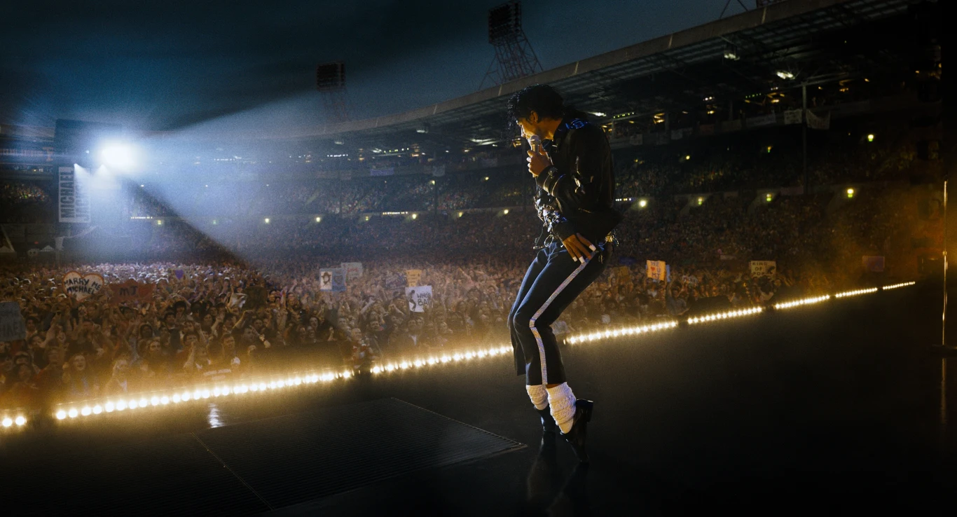 Kadr z filmu "Michael" Wokalista w czarno-białym stroju wykonuje charakterystyczny taniec na oświetlonej scenie, skierowany do ogromnej, entuzjastycznej publiczności na stadionie, z wyraźnym snopem światła skierowanym z boku.
