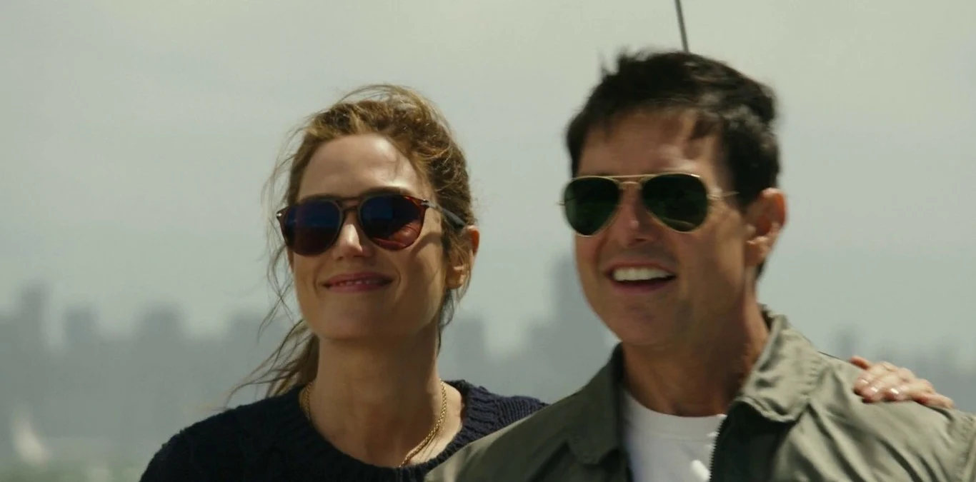 "Top Gun: Maverick" Dwoje ludzi w okularach przeciwsłonecznych; kobieta z włosami związanymi, mężczyzna w lotniczej kurtce, w tle miasto.