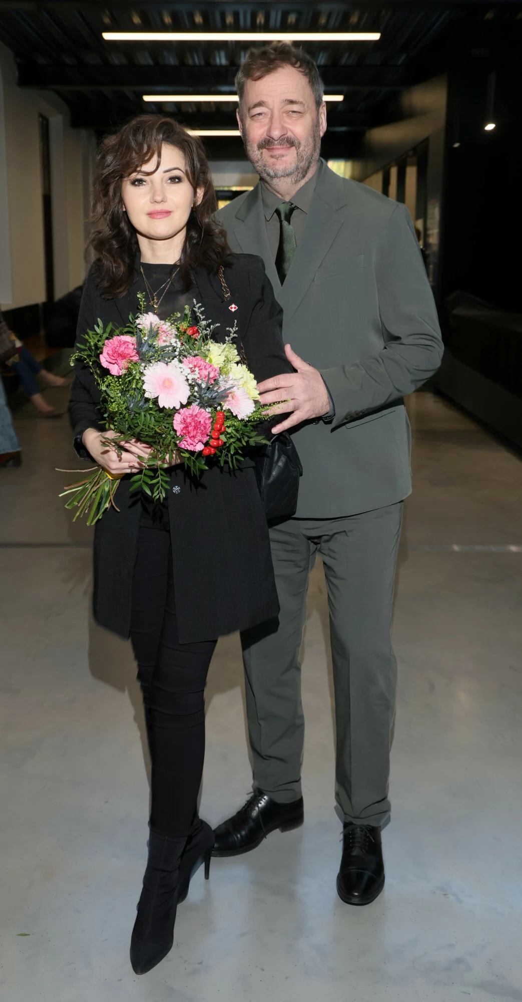 Kobieta w czarnym ubraniu trzymająca bukiet kolorowych kwiatów stoi obok mężczyzny w zielonym garniturze, oboje uśmiechnięci, sfotografowani w nowoczesnym budynku o jasnym wnętrzu.