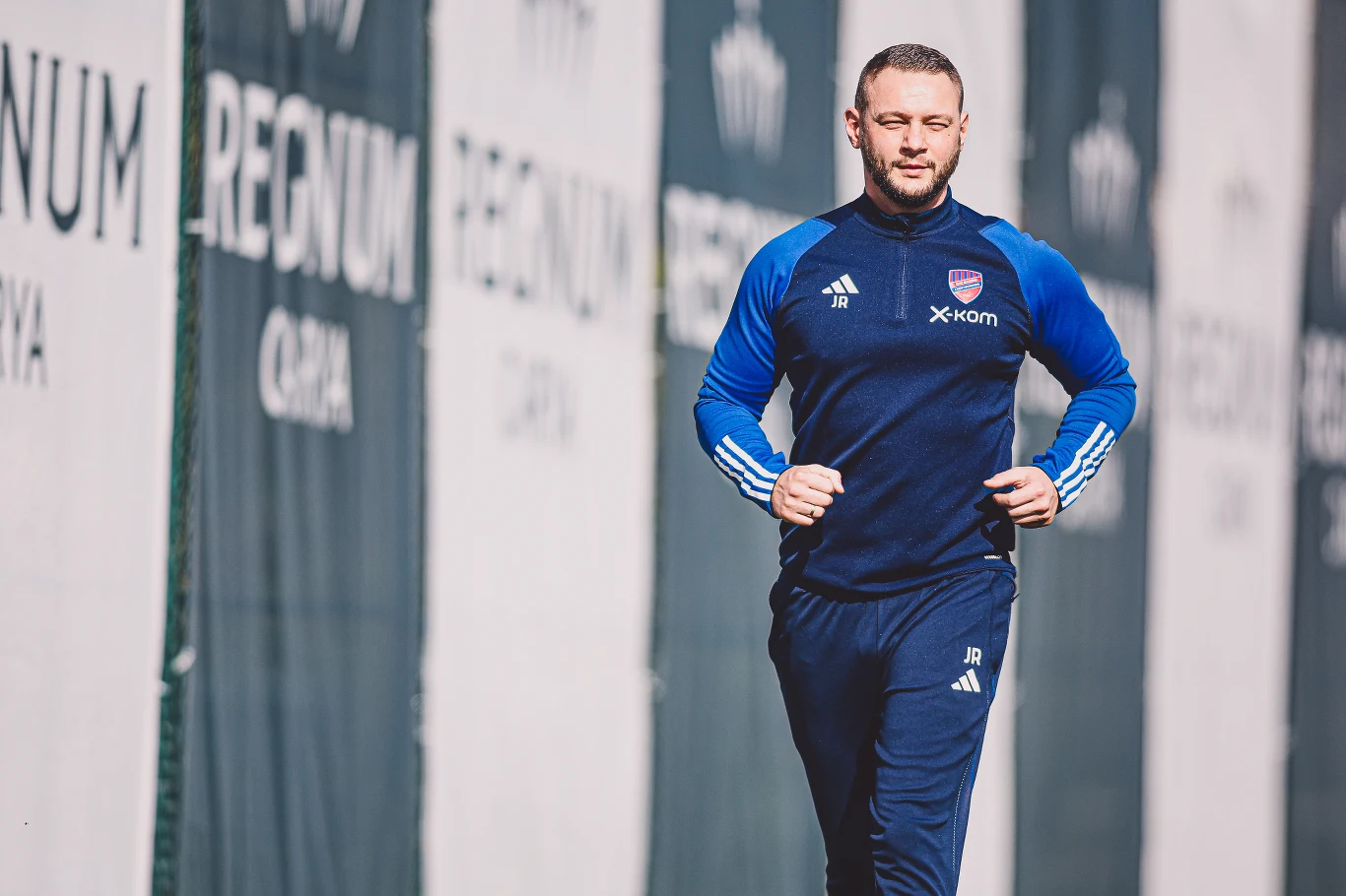 Jakub Rajek, kitman Rakowa Częstochowa mężczyzna w sportowym stroju w barwach niebiesko-granatowych biegnie obok ogrodzenia z powtarzającym się napisem REGNUM