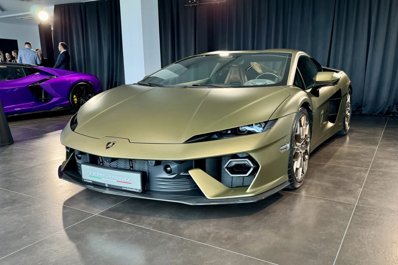 matowozielony Lamborghini z nisko osadzonym przodem i agresywnymi wlotami powietrza, stojący w jasnym, nowoczesnym salonie na tle ciemnych zasłon; w tle widoczny fioletowy supersamochód