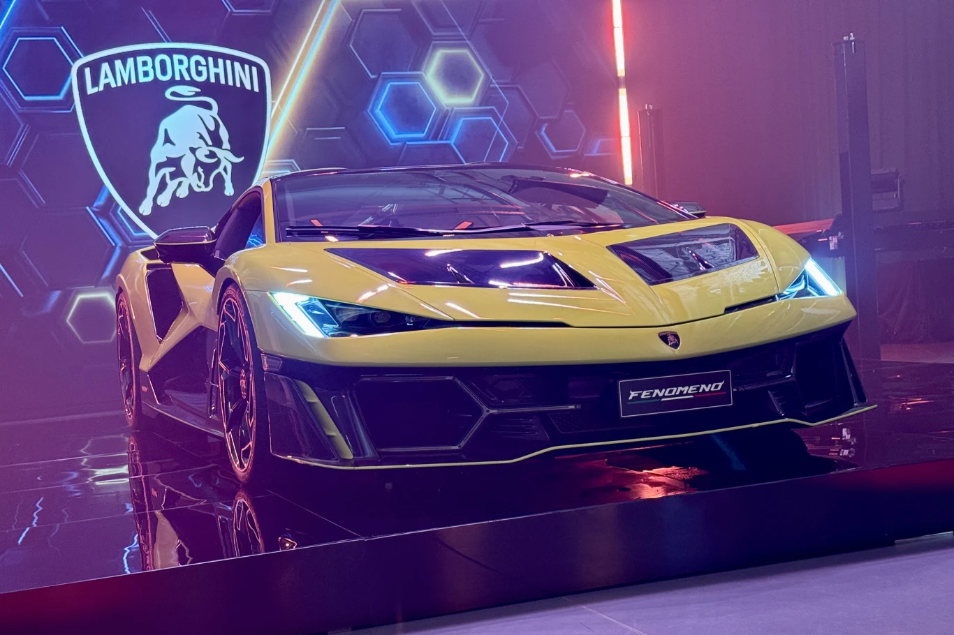 Jaskrawożółty Lamborghini z charakterystycznymi ostrymi liniami nadwozia stoi na podeście wystawowym, z tyłu widoczne logo marki oraz dynamiczne oświetlenie podkreślające sportowy charakter auta.