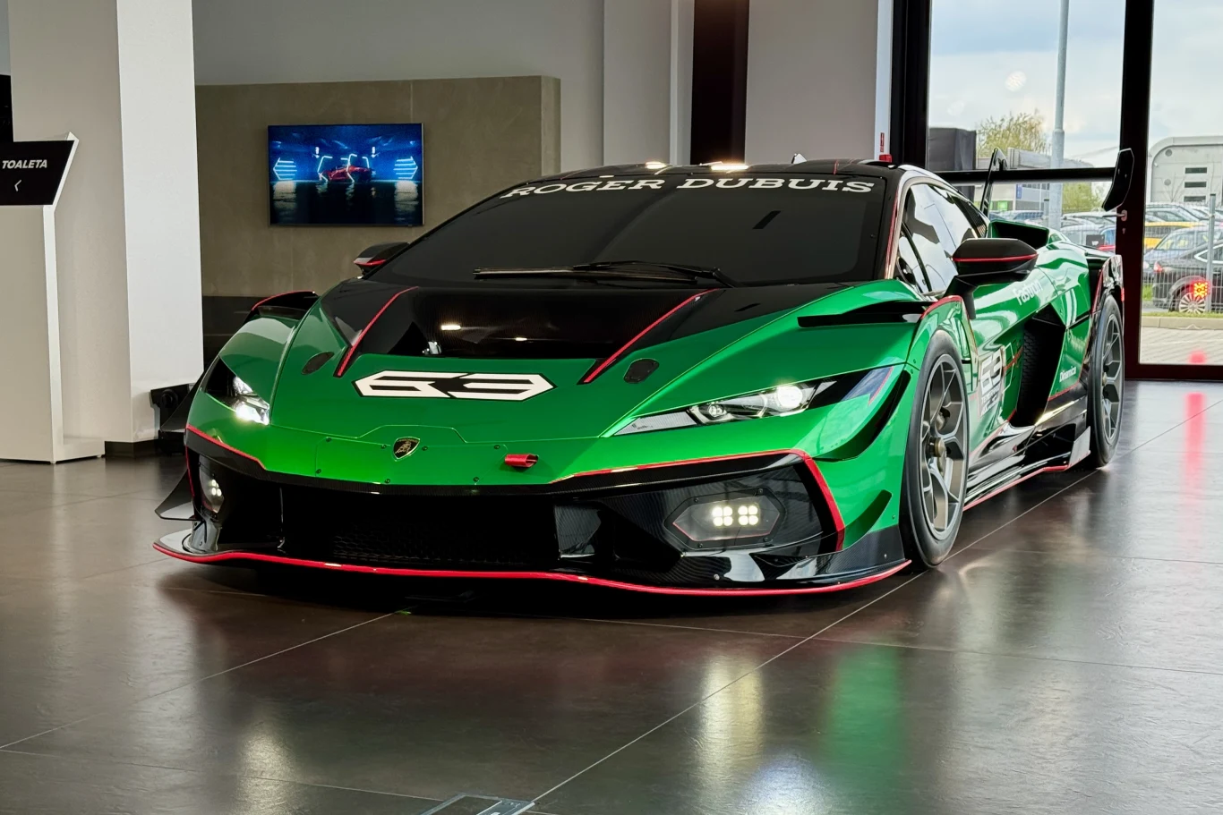 zielone, sportowe Lamborghini z agresywnym aerodynamicznym pakietem, umieszczone w nowoczesnym wnętrzu salonu motoryzacyjnego; samochód wyróżnia się niską sylwetką, szerokimi nadkolami i dużym tylnym skrzydłem, na szybie widoczny biały napis 'Roger Dub...