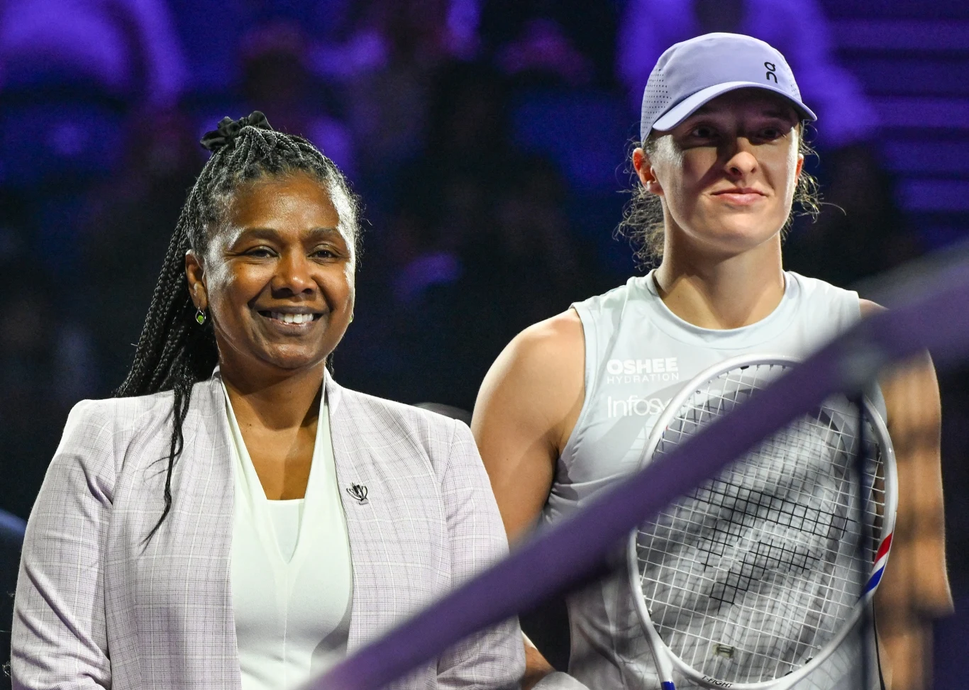 Portia Archer i Iga Świątek dwie kobiety stoją obok siebie na tle ciemnego, oświetlonego stadionu; jedna z nich ubrana w jasny sportowy strój i czapkę, trzyma rakietę tenisową, druga ubrana w jasny garnitur uśmiecha się do obiektywu