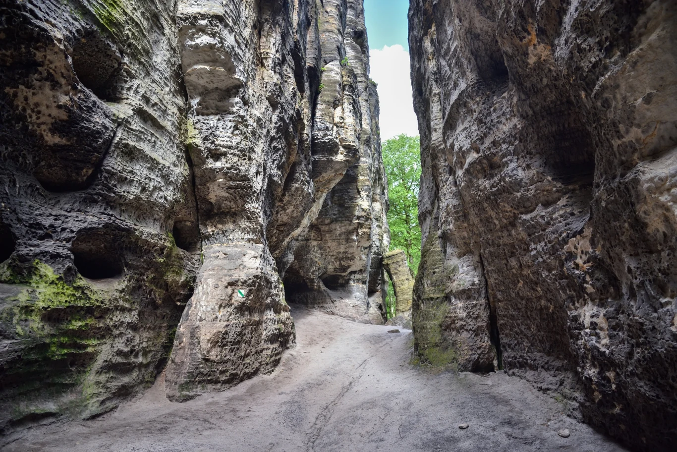 Wąska ścieżka prowadzi pomiędzy wysokimi, pionowymi skałami z licznymi wgłębieniami i nierównościami, na końcu prześwituje zielone drzewo oraz niebo.