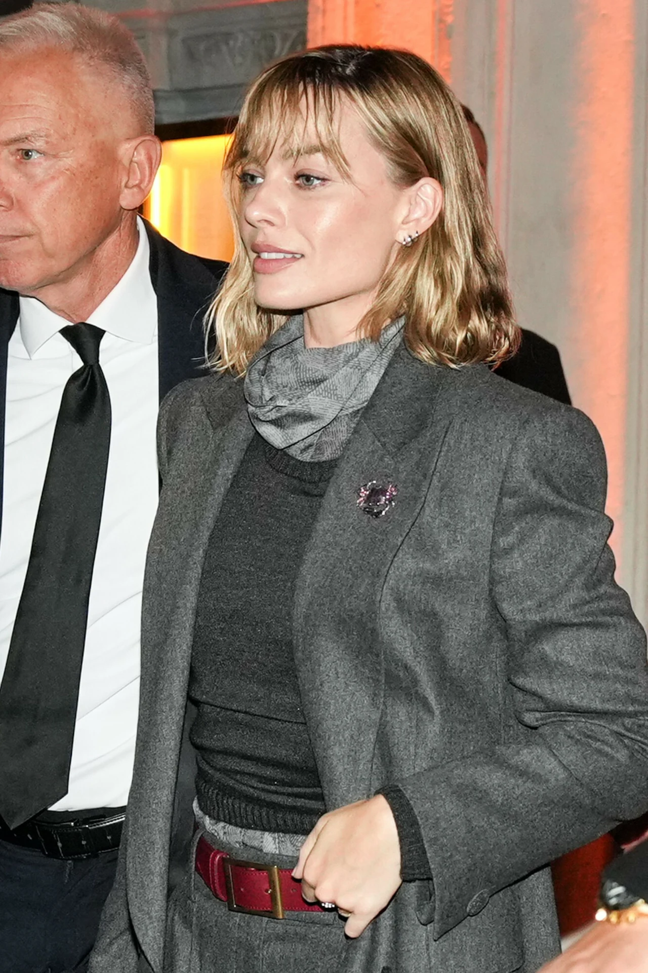 Margot Robbie pokazuje, że jest niczym kameleon w świecie mody. Jej różne "wcielenia" bywają hipnotyzujące Kobieta w eleganckim szarym garniturze z broszką na klapie i szarym szalem na szyi, z krótkimi blond włosami i naturalnym makijażem, stoi obok mężczyzny w czarnym garniturze i białej koszuli z czarnym krawatem; tło z ciepłym, pomarańczowym światłem.