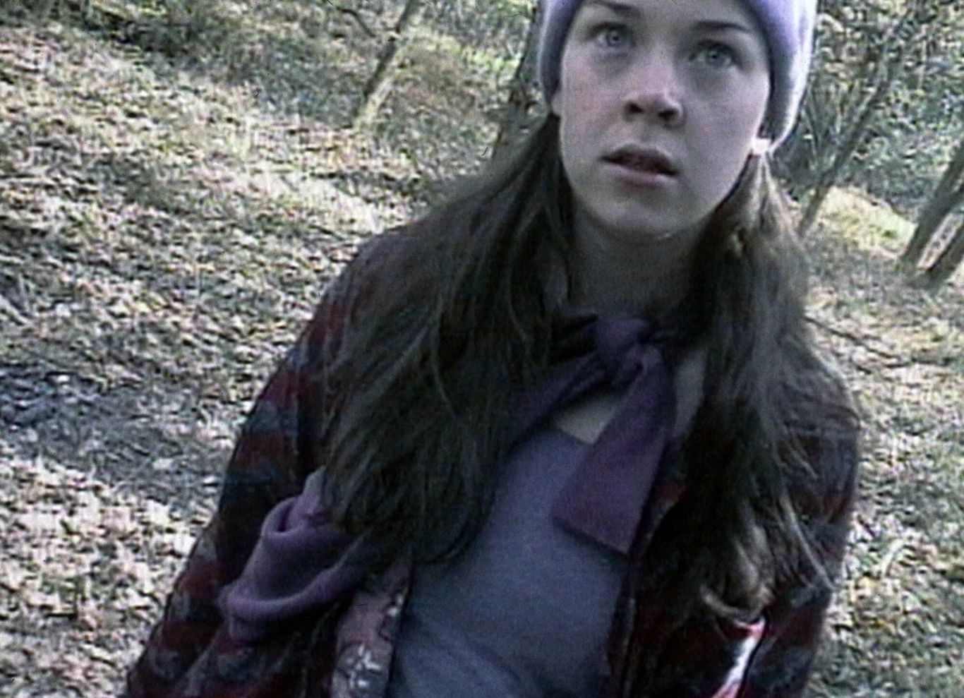 "The Blair Witch Project" Młoda kobieta o długich, ciemnych włosach, ubrana w fioletową czapkę i sweter, stoi w lesie patrząc przed siebie z poważnym wyrazem twarzy; wokół niej widoczne drzewa i opadłe liście na ziemi.