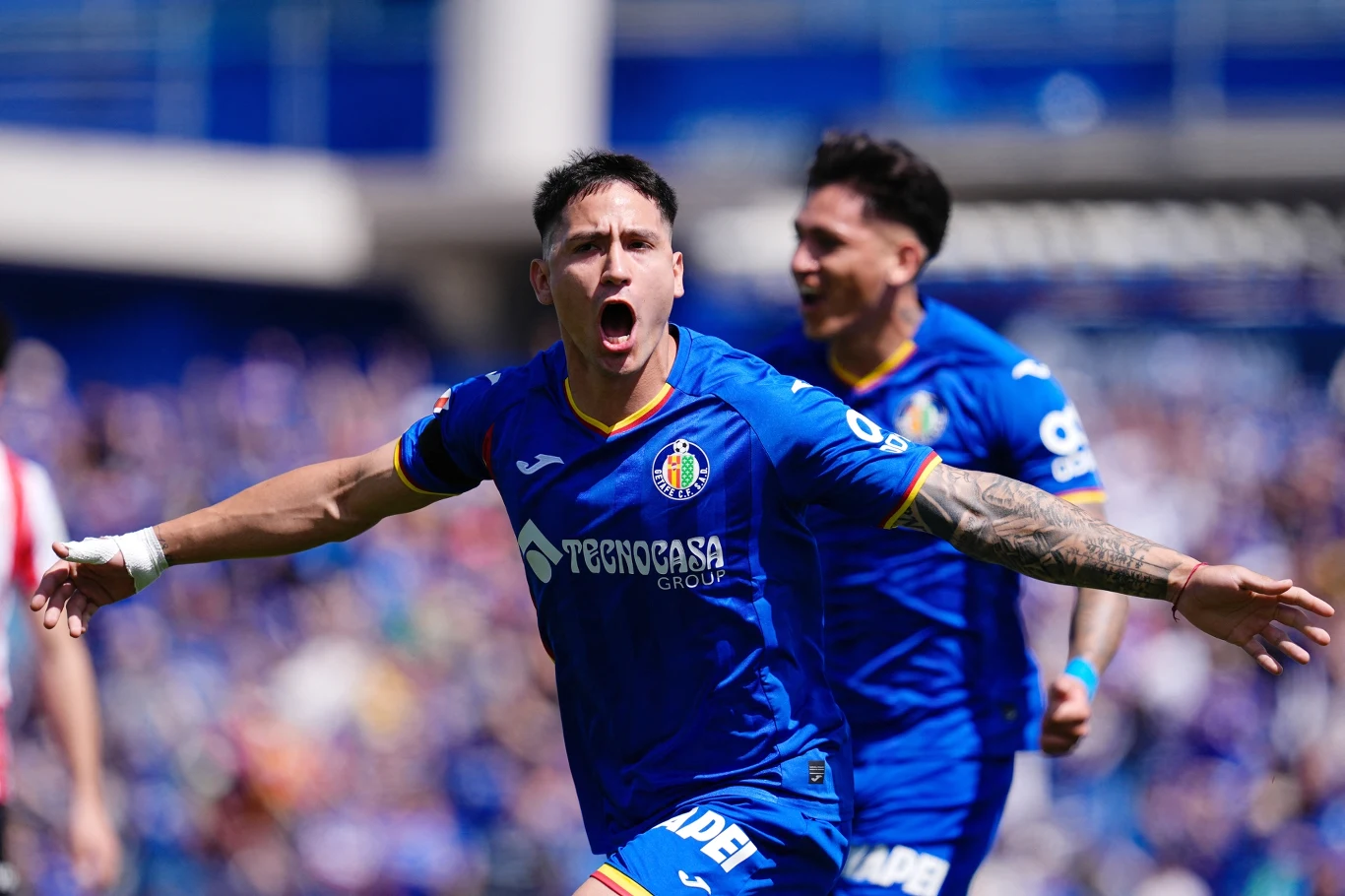 piłkarz w niebieskim stroju z logo Getafe CF świętuje zdobycie gola, rozkładając szeroko ramiona, a w tle drugi zawodnik z tej samej drużyny cieszy się razem z nim na boisku podczas meczu