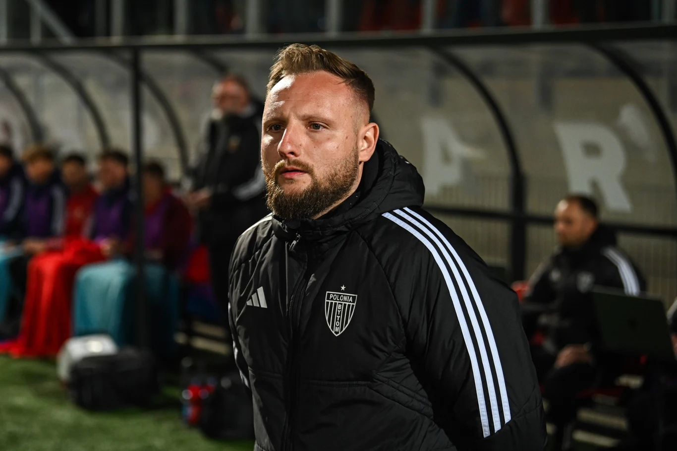 trener w sportowej kurtce Adidas z herbem klubu Aston, stojący przy linii bocznej boiska, w tle ławka rezerwowych z grupą zawodników