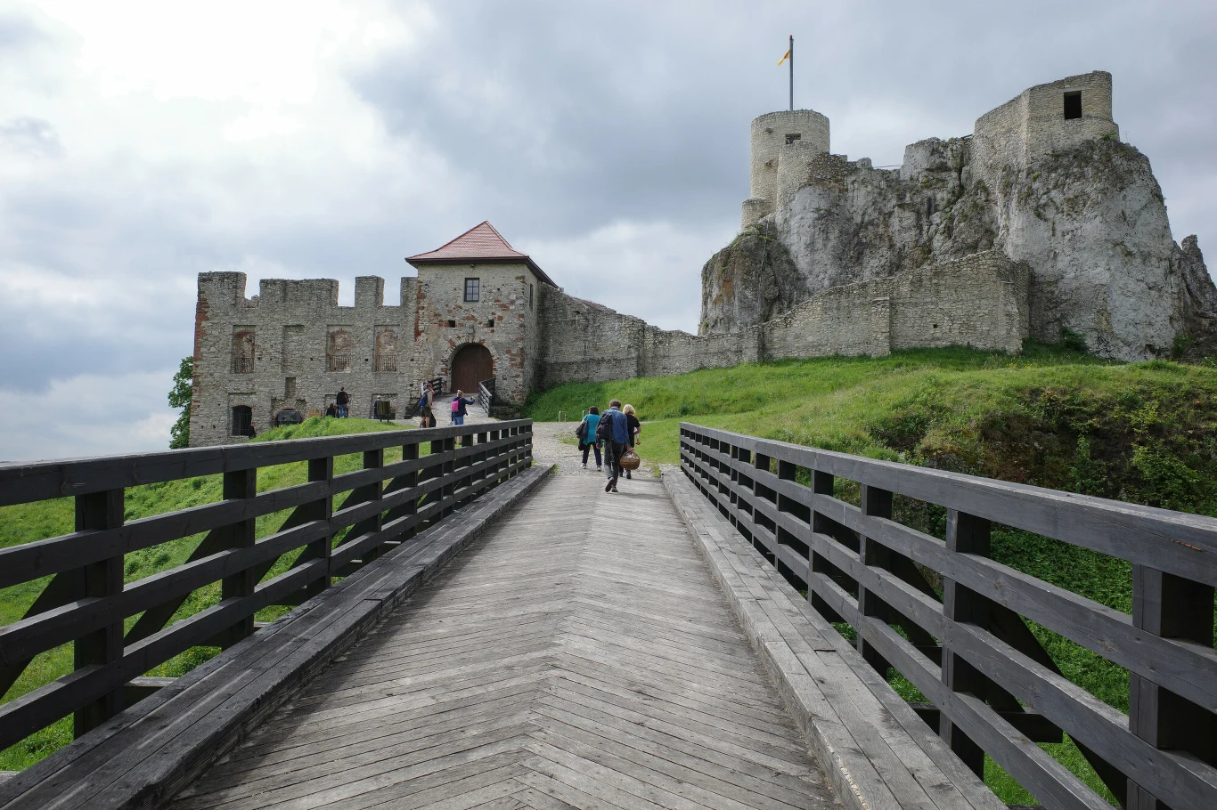 Przy zamku w Rabsztynie powstaną nowe atrakcje dla turystów Kamienny zamek w Rabsztynie z okrągłą wieżą, mostem i grupą osób spacerujących otoczony zieloną trawą