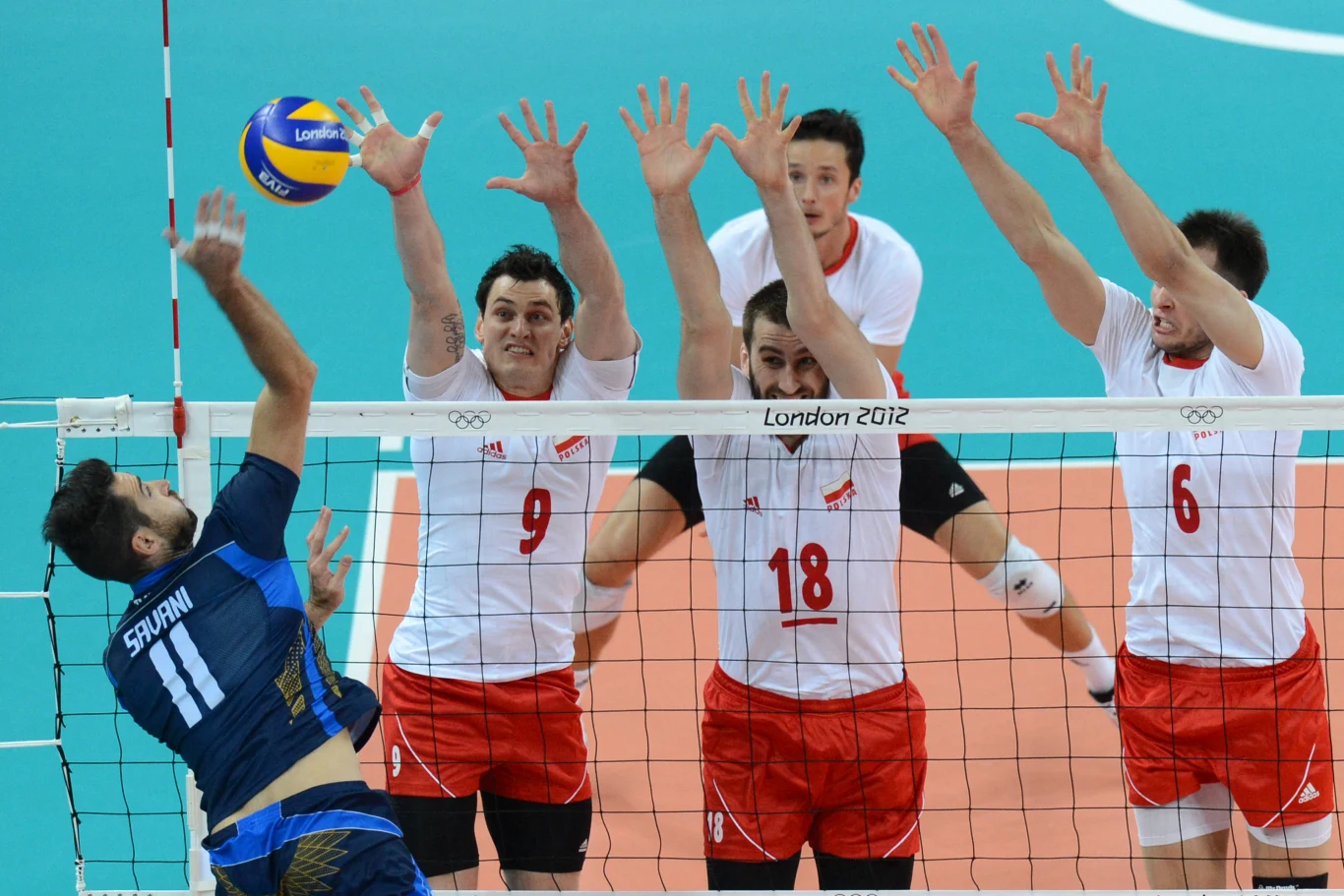 zawodnik w niebieskim stroju atakuje piłkę siatkową, naprzeciwko trzech graczy w biało-czerwonych strojach ustawionych do bloku przy siatce w trakcie meczu olimpijskiego