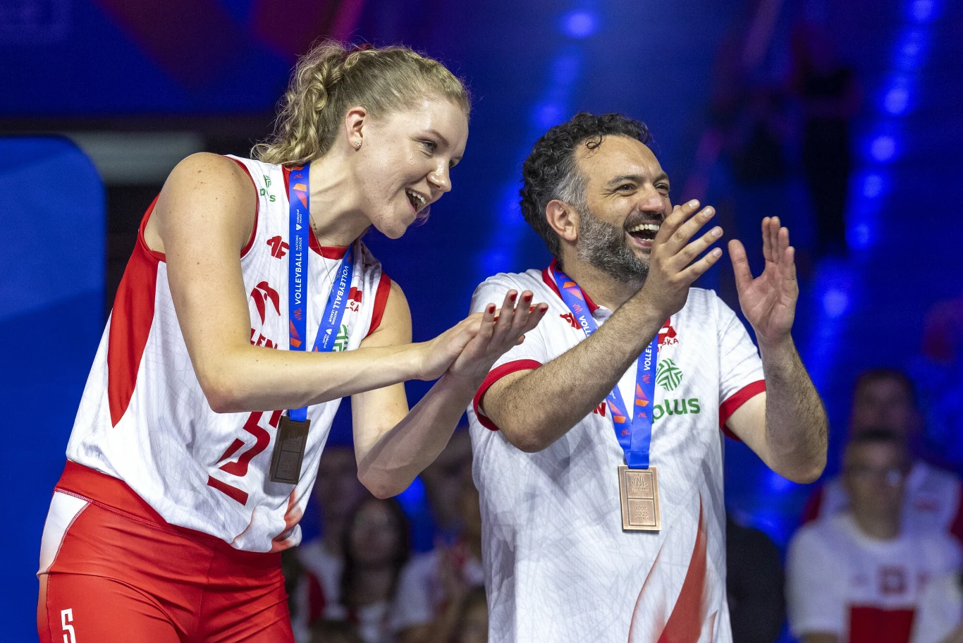 zawodniczka i trener polskiej drużyny siatkówki w sportowych strojach, uśmiechnięci, z medalami na szyi, cieszą się z sukcesu podczas ceremonii wręczenia nagród