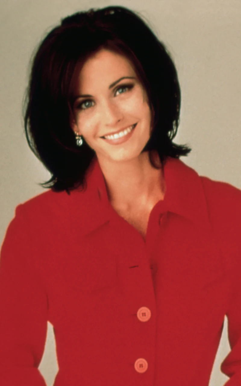 kobieta z ciemnymi włosami i jasną cerą uśmiecha się, ubrana w elegancki czerwony płaszcz z dużymi guzikami i delikatne kolczyki, tło jednolite i jasne